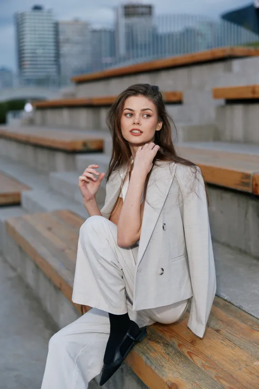 woman in light beige suit sitting on urban wooden stairs with thoughtful expression, fashion style, casual business outfit, spring mood, city background