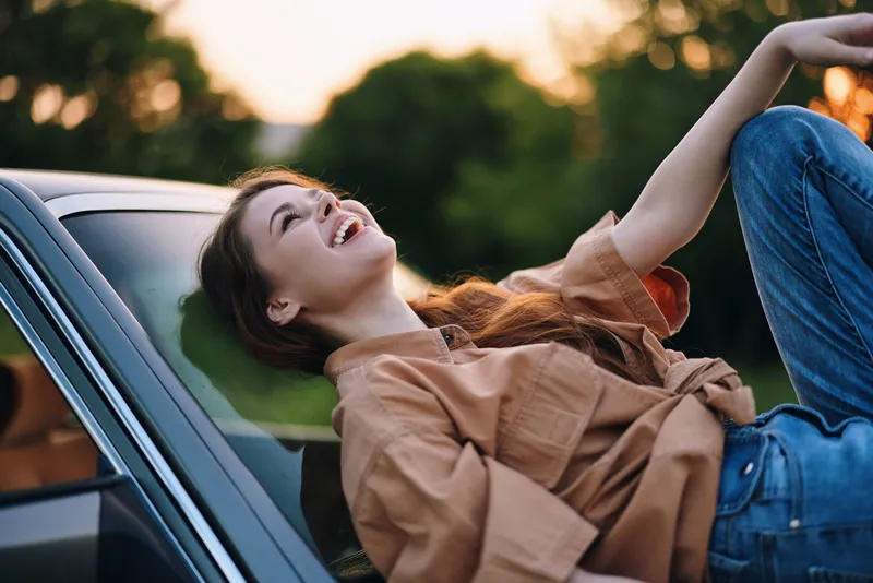 Young woman laughing joyfully on a car hood during sunset, wearing a casual brown shirt and blue jeans, showcasing a carefree lifestyle in nature.