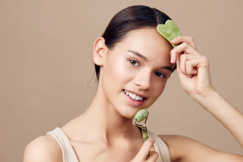 Young woman applying facial massage with jade tools for skincare, promoting beauty and wellness in a minimalist setting