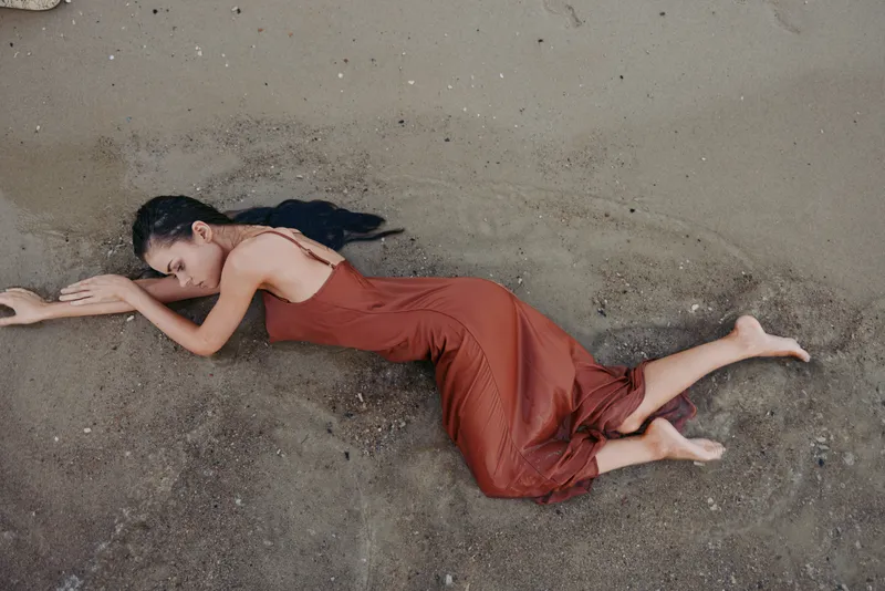 A female figure lies prone on wet sand wearing a flowing red dress, posture stretched and quiet, evoking fatigue, vulnerability, and silent introspection.