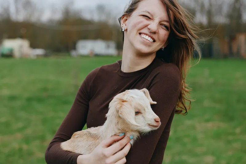 A smiling woman in a brown sweater holds a young goat kid in a sunny rural field, capturing a candid lifestyle moment of affection and companionship with animals.