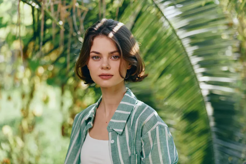 A young woman stands outdoors among lush greenery and palm fronds, wearing a striped green jacket over a white top, with a calm, confident gaze.