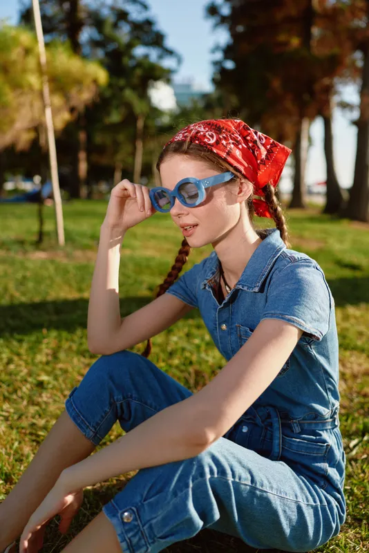 Young woman with braids sitting on grass wearing blue denim outfit and red bandana enjoying sunny park outdoors in casual summer style.
