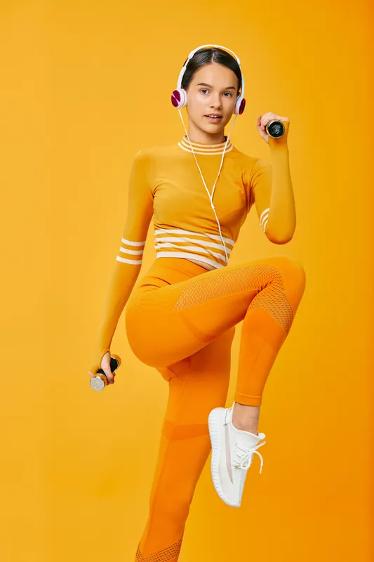 Young woman fitness model performing a high knee exercise with dumbbells and headphones in a monochrome orange studio. Sport outfit, white sneakers, confident focused pose.