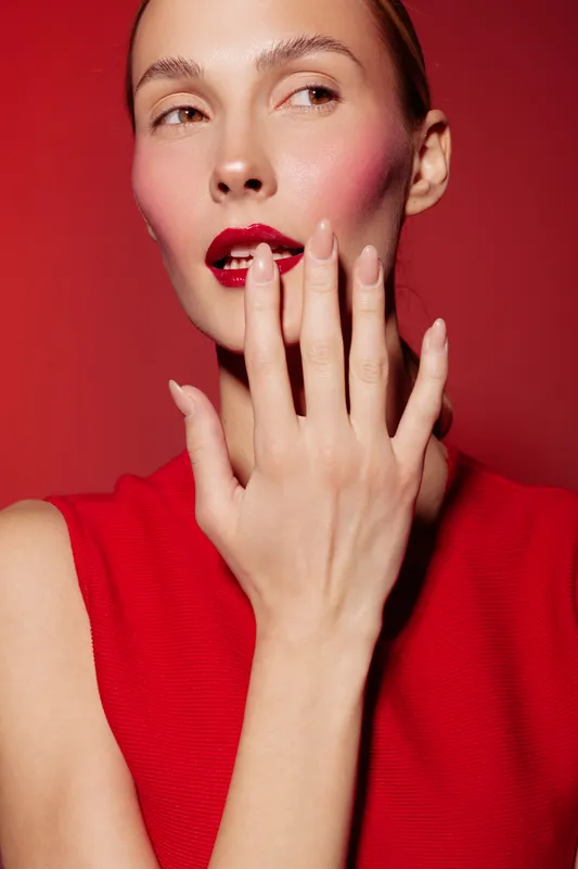Close-up of a woman with red lipstick and blush wearing a red top, touching her lips with manicured fingers. Concept of beauty, makeup, fashion, and elegance on red background.