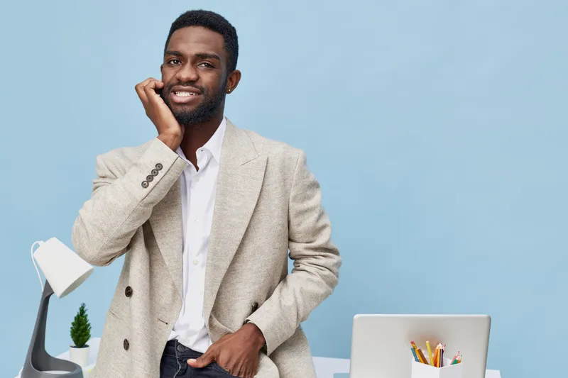 Happy confident man with a beard, dressed in a beige blazer and white shirt, posing with a smile on his face in front of a light blue background. Studio portrait, casual business style, positive mood,