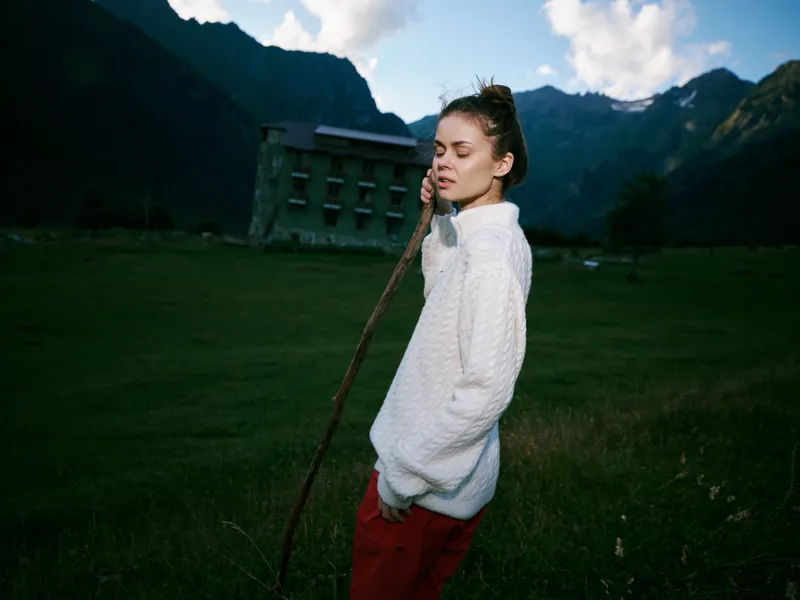 Woman in a white sweater holds a stick in an open field with distant mountains, showcasing outdoor lifestyle, natural scenery and rural landscape for travel and adventure visuals