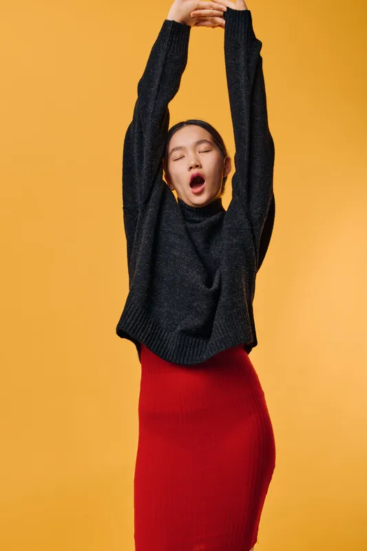 Young woman stretching with eyes closed wearing black sweater and red skirt on yellow background. Casual outfit, relaxed mood and breathing deep for wellness and comfort.