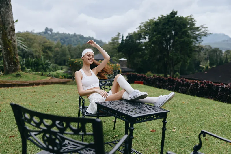 Young woman enjoying a peaceful moment outdoors, wearing a white outfit and headband, surrounded by greenery and flowers, smiling joyfully in a serene setting