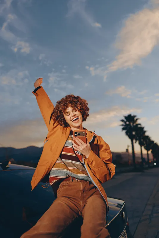 Woman lifestyle selfie with smartphone outdoors at sunset, palm trees street casual jacket, curly haired woman seated on car hood, smiling, warm cinematic light