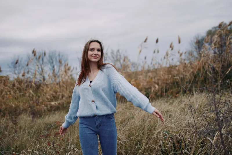 A young woman in a cozy cardigan strolls through tall grass, captured in natural light with a genuine expression and documentary approach that conveys authenticity and reliability.