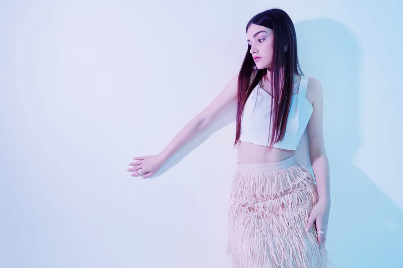 Young woman in white top and fringe skirt poses against a light wall. Her long dark hair frames her face as she looks to the side. Concept of beauty fashion and style.