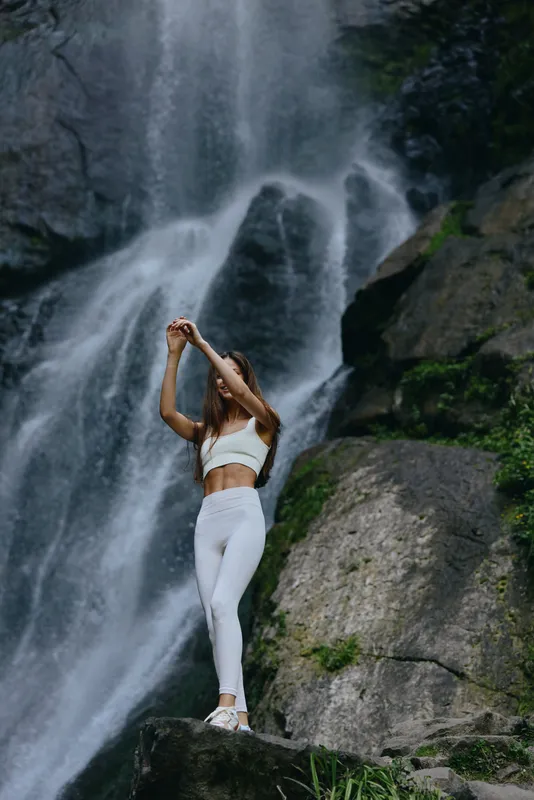 woman practicing yoga by waterfall in white activewear, outdoor meditation, fitness, nature, healthy lifestyle, serene atmosphere