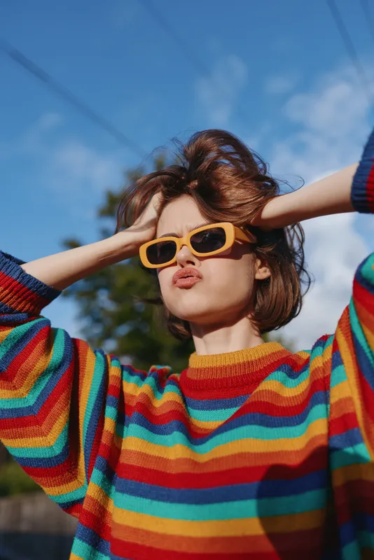 Young woman in a striped sweater and yellow sunglasses, outdoor portrait showing casual fashion, playful pose and short brown hair under sunny sky, puckered lips and hands in hair.