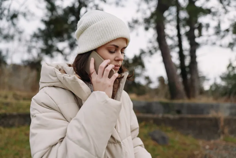 Woman on a phone outdoors in winter, wearing a thick coat and beanie, talking amid a chilly setting with trees and a quiet landscape, conveying communication and everyday outdoor activity