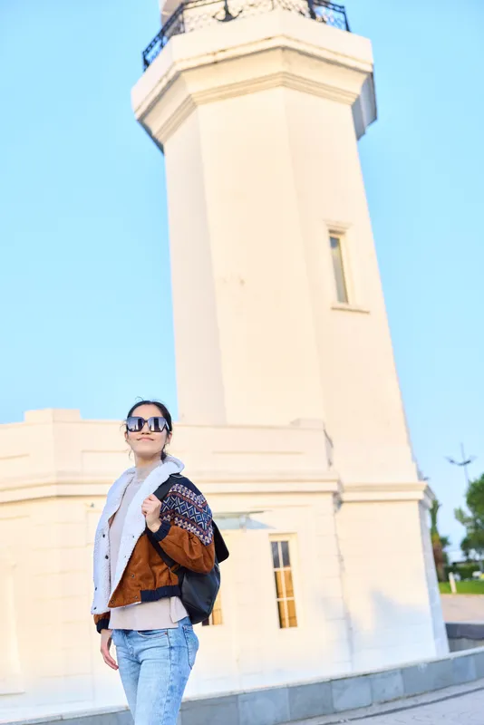 Woman in casual fashion with sunglasses stands beside a tall lighthouse on a sunny day, showcasing travel style and urban exploration photography vibes