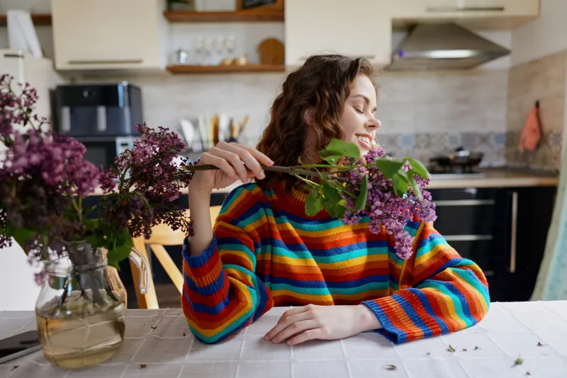 Woman in a colorful striped sweater sits at home in a bright kitchen, admiring a bouquet of purple flowers, a calm cozy domestic moment of everyday life