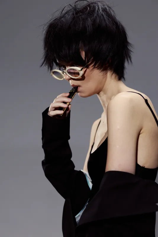 Stylish woman with short black hair and glasses wearing a dark jacket and black lingerie posing with a thoughtful expression