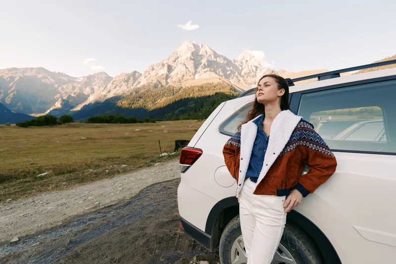 Woman by car on a mountain road landscape, travel scene with warm jacket outdoors beside an SUV on a dirt track, relaxed pose and panoramic alpine view at golden hour.