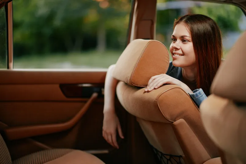 Young woman in casual attire smiling and leaning out of a car window, surrounded by greenery, evoking a sense of adventure and joy in summer.
