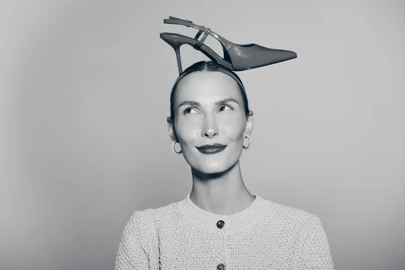 Young caucasian woman with pale skin and short hair holds high heel shoe on head, wearing light sweater, smiling.