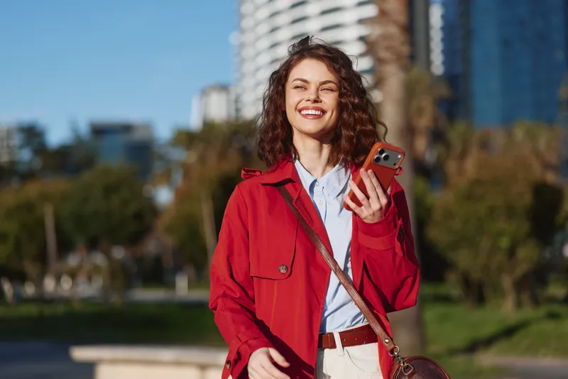 Smiling woman with curly hair wearing a red jacket holds a smartphone in a sunny city park, bright background showing modern buildings and blue sky.