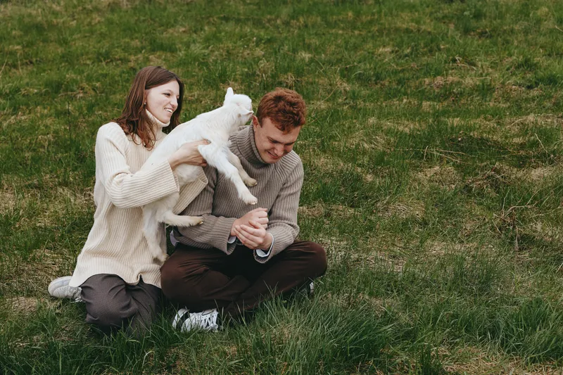 Young couple enjoys playful moment with a white baby goat while sitting on green grass in casual sweaters during a relaxed lifestyle outdoor scene.