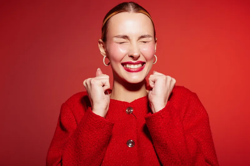 Excited young woman in red sweater smiles with closed eyes and clenched fists on red background. Concept of happiness, joy, celebration, and positive emotions in studio lighting.