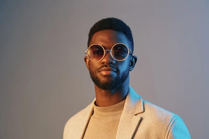 Stylish young man in unique round sunglasses and tailored blazer against a gradient background, showcasing contemporary fashion and confidence