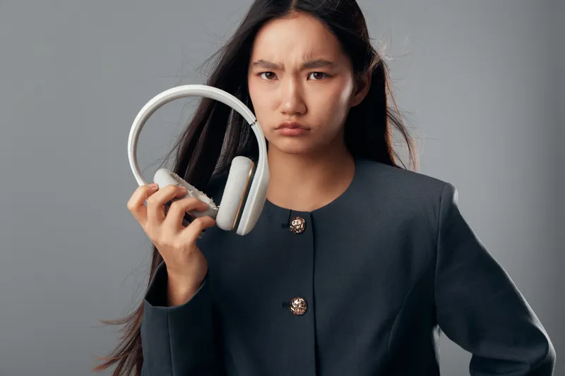 Young woman with long hair wearing dark jacket holds white headphones with serious facial expression on gray background. Portrait of female with audio device in hand and focused look.