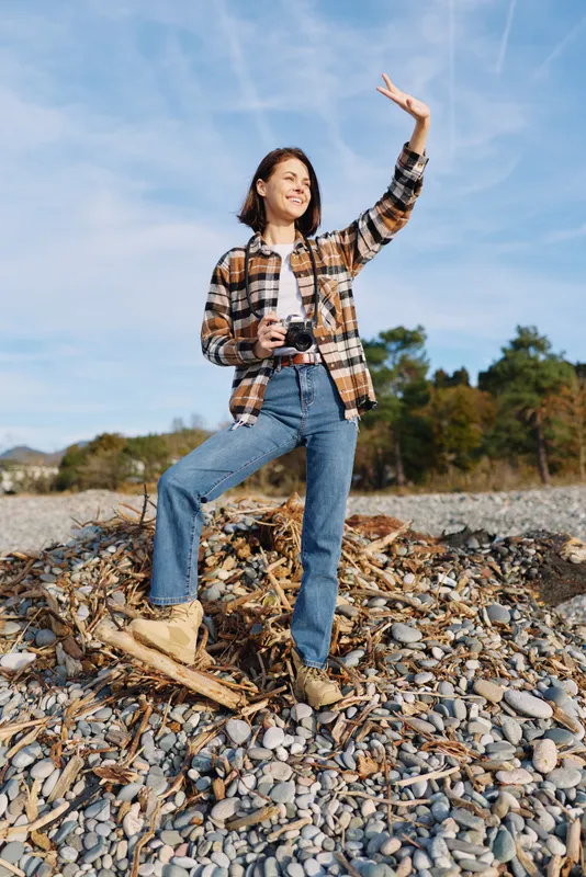 Woman standing on a rocky beach with a camera in hand, wearing a plaid shirt and jeans, raising her arm against the blue sky, conveying outdoor exploration and casual adventure.