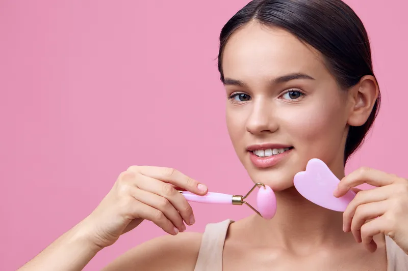 Young woman with a pink roller tool for skincare, smiling against a soft pink background, embodying beauty and self care concepts
