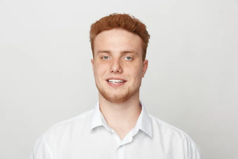 Smiling young man with red hair and freckles wears white shirt, looking at camera on light background. Portrait of confident male model with natural expression and casual style.