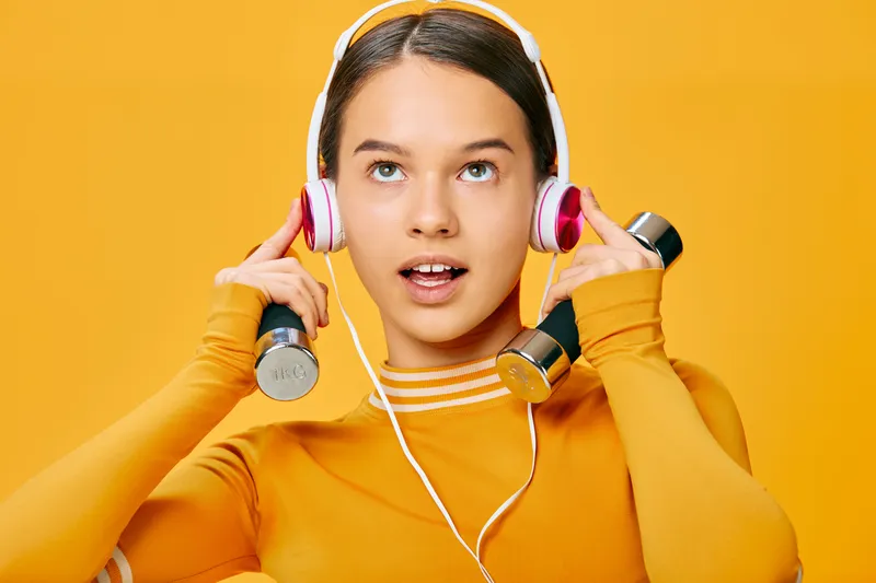 Woman in an orange athletic top lifts dumbbells with headphones on, bright studio backdrop, focused on workout and energy for fitness and sport motivation during training session.