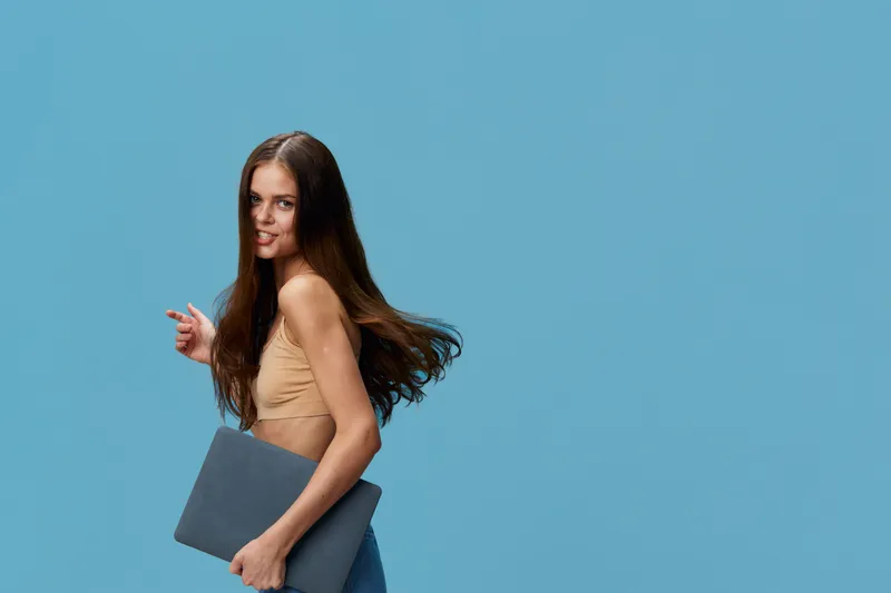 young woman holding laptop and smiling, long brown hair, casual beige top against blue background, bright and confident atmosphere, modern lifestyle