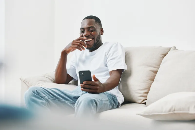 Happy young African American man laughing while using a smartphone at home, wearing casual white t shirt and jeans in a bright, minimalistic living room
