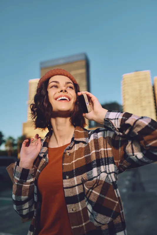 woman smiling phone urban candid golden hour authenticity lifestyle mindful living emotional storytelling warm glow as she talks outdoors against city skyline in sunset light