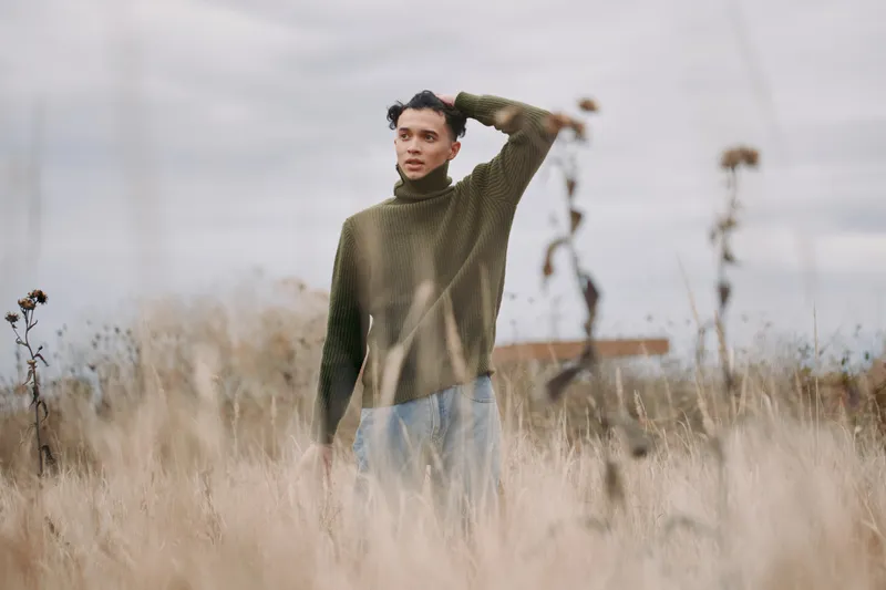 A solitary young man in a textured green sweater stands amid tall dry grass, captured in an authentic, natural portrait that emphasizes genuine expression and understated realism, with quiet
