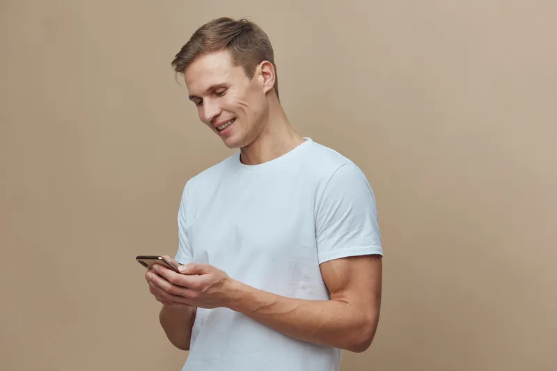 Smiling young man in casual white t-shirt using mobile phone, cheerful expression, engaging with device, casual indoor setting, beige background, lifestyle, everyday life, modern technology concept