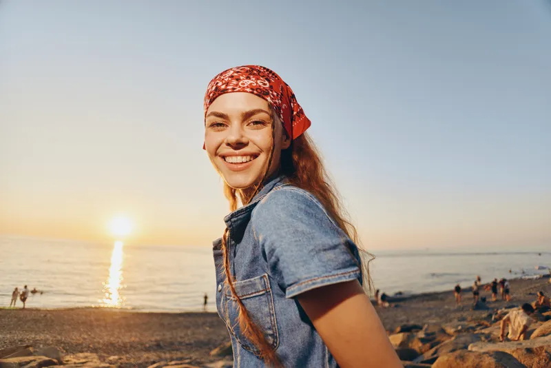 Lifestyle woman in modern boho-western denim street style smiling outdoors at sunset with warm film color and casual summer vibe on the beach