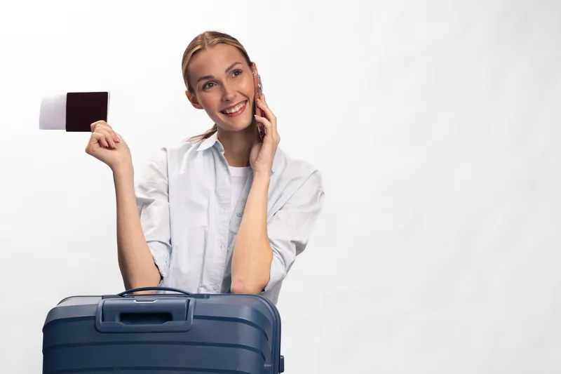 Smiling young woman with suitcase holds passport and talks on phone, ready for travel. White background emphasizes her happy and confident mood before trip.