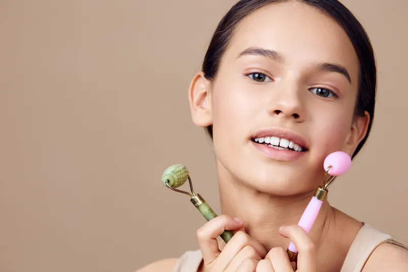 Young girl with natural makeup holding facial rollers, showcasing a glowing complexion against a neutral backdrop focused on skincare and beauty