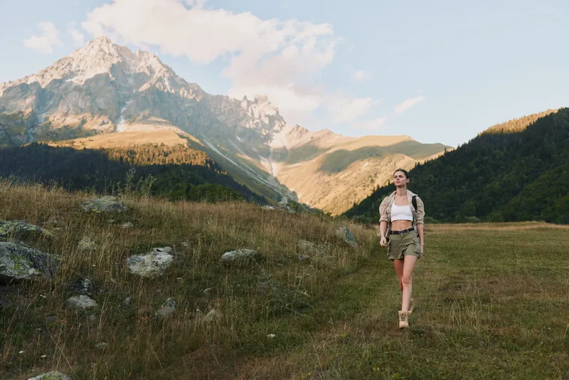 Young woman with long hair walks confidently through a mountain meadow wearing a white crop top and khaki shorts during a sunny day.