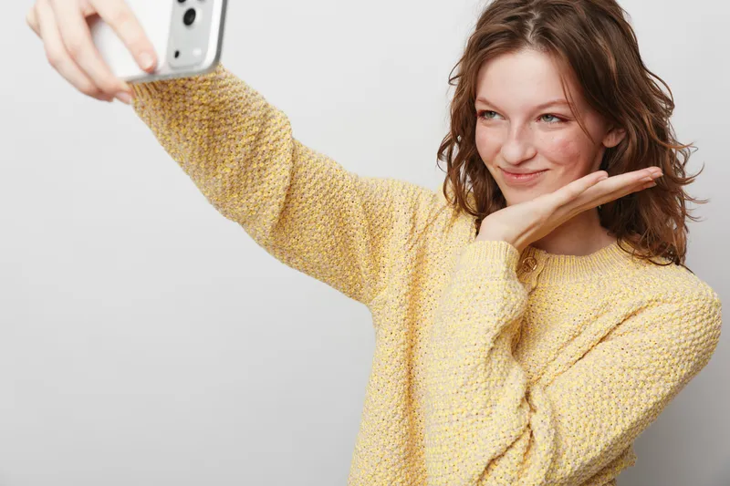 Young woman in yellow sweater takes a selfie with smartphone, smiling and posing with hand near face. Casual lifestyle, technology, and social media concept on white background.