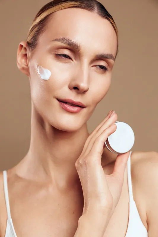 Woman skincare face cream beauty spa cotton pad hygiene treatment, applying cream with cotton pad on face, smooth clean skin, studio beige background, calm expression