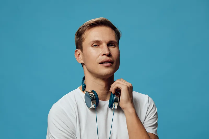 Male DJ wearing white t-shirt and headphones around neck pose against plain blue background. People, music, entertainment concept, lifestyle, casual style, modern, young man.