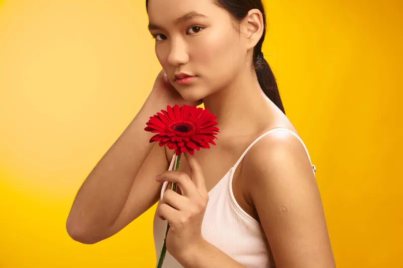 Asian woman holds a bright red flower in a close portrait, set against a warm yellow backdrop, conveying calm elegance and modern beauty in a studio style.