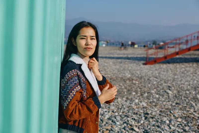 Woman leaning against a turquoise wall on a pebbled beach, wearing a patterned jacket and warm fleece collar, capturing a relaxed outdoor mood and coastal scene