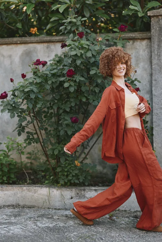 A stylish woman enjoys an outdoor stroll in an orange suit, curly hair, and a bright smile, portraying confident lifestyle and casual fashion amid blooming roses