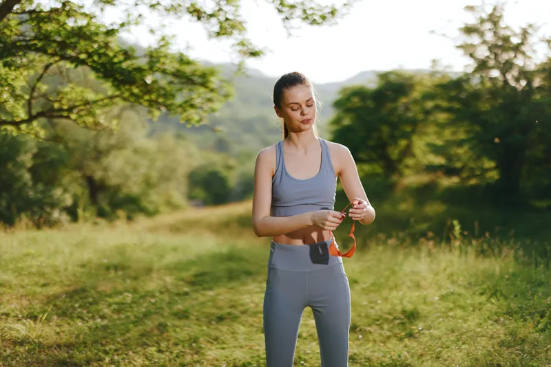 Young woman in activewear standing on a sunny trail, adjusting fitness tracker with a determined expression, surrounded by lush green nature